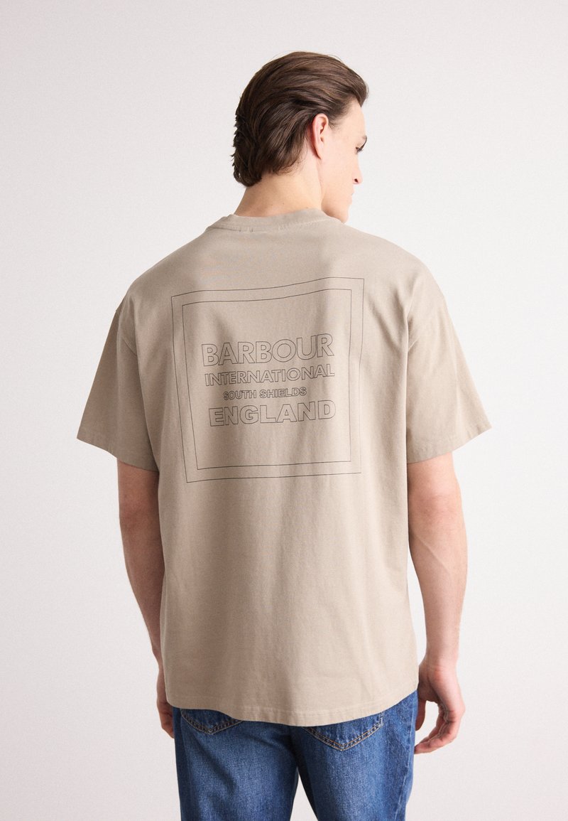 Tricou din bumbac bej, cu un grafic dreptunghiular în cerneală închisă pe spate, cu textul: "BARBOUR INTERNATIONAL SOUTH SHIELDS ENGLAND."