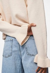 Jasnoróżowy sweter z dzianiny z prążkowanymi mankietami, noszony na wysokich niebieskich dżinsach. Sweter ma luźny krój i miękką teksturę.