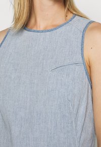 Ljusblå ärmlös topp av denimtyg med en slät textur. Har en liten bröstficka och kontrasterande sömnad längs kanterna.