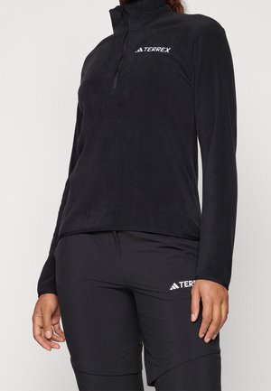 Personne portant un haut noir à manches longues avec une fermeture éclair à mi-longueur et un pantalon assorti, tous deux arborant des logos Terrex blancs sur la poitrine et la cuisse.