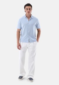 Camicia azzurro chiaro a maniche corte con una texture morbida, abbinata a pantaloni bianchi larghi e scarpe slip-on grigie.