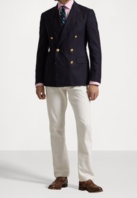 Blazer doppiopetto blu navy con bottoni dorati, camicia rosa, cravatta a fantasia verde e blu, pantaloni avorio e scarpe eleganti marroni.