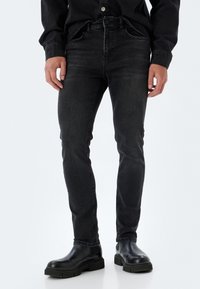 Jeans de mezclilla negros con un ajuste Slim, que cuentan con cinco bolsillos, lazos para cinturón y un ligero desgastado. Llevados con botas negras hasta el tobillo.