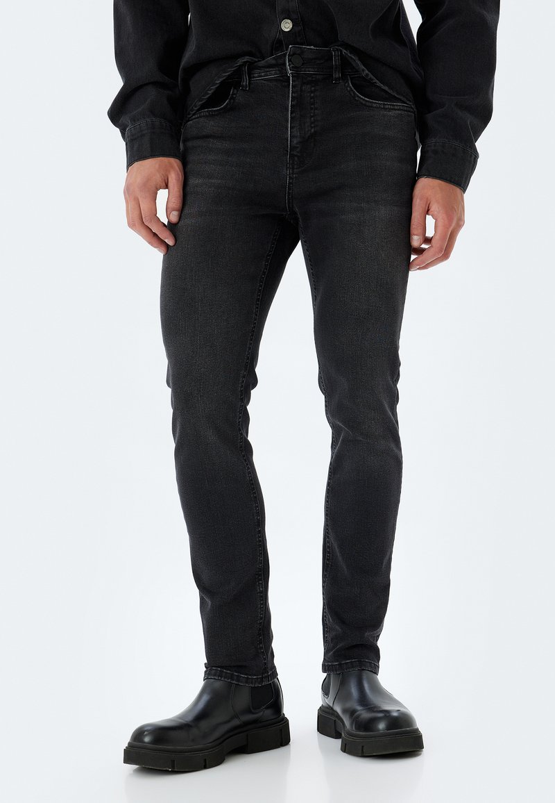 Jeans de mezclilla negros con un ajuste Slim, que cuentan con cinco bolsillos, lazos para cinturón y un ligero desgastado. Llevados con botas negras hasta el tobillo.