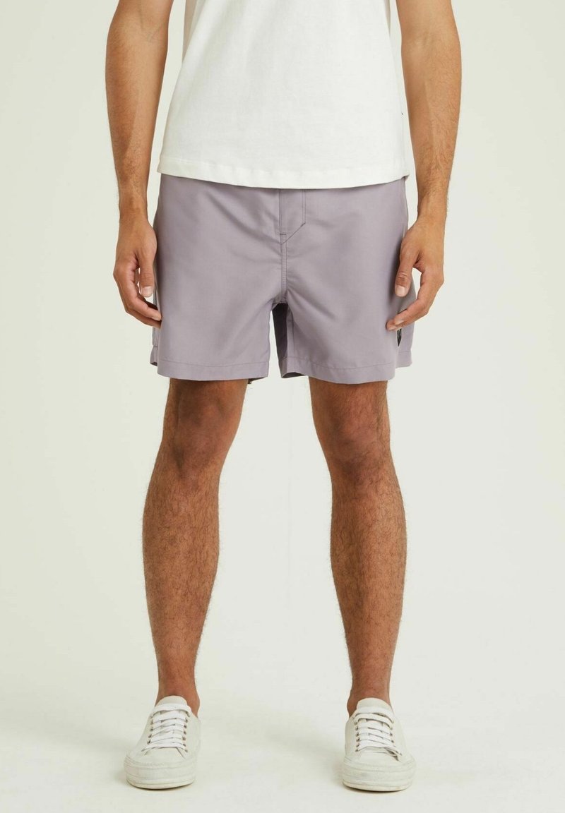 Homme portant un short gris clair, des baskets blanches et un t-shirt blanc, debout devant un fond clair uni.
