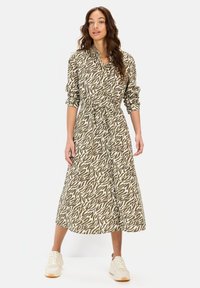 Langsleeve, button-down jurk in olijfgroen en crème abstract patroon. Heeft een tailleband en valt tot halverwege de kuit, gecombineerd met witte sneakers.