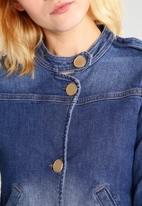 Sininen farkkutakki, jossa on korkea kaulus, kultaiset nepparit ja kaksi etutaskua. Hieman vaaleampi sävy helman kohdalla.