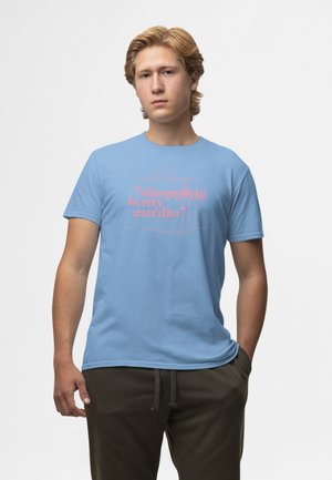 Jonge man met blond haar die een lichtblauw T-shirt draagt met de roze tekst "shoppen is mijn cardio" en een omtrek van een winkelzak.