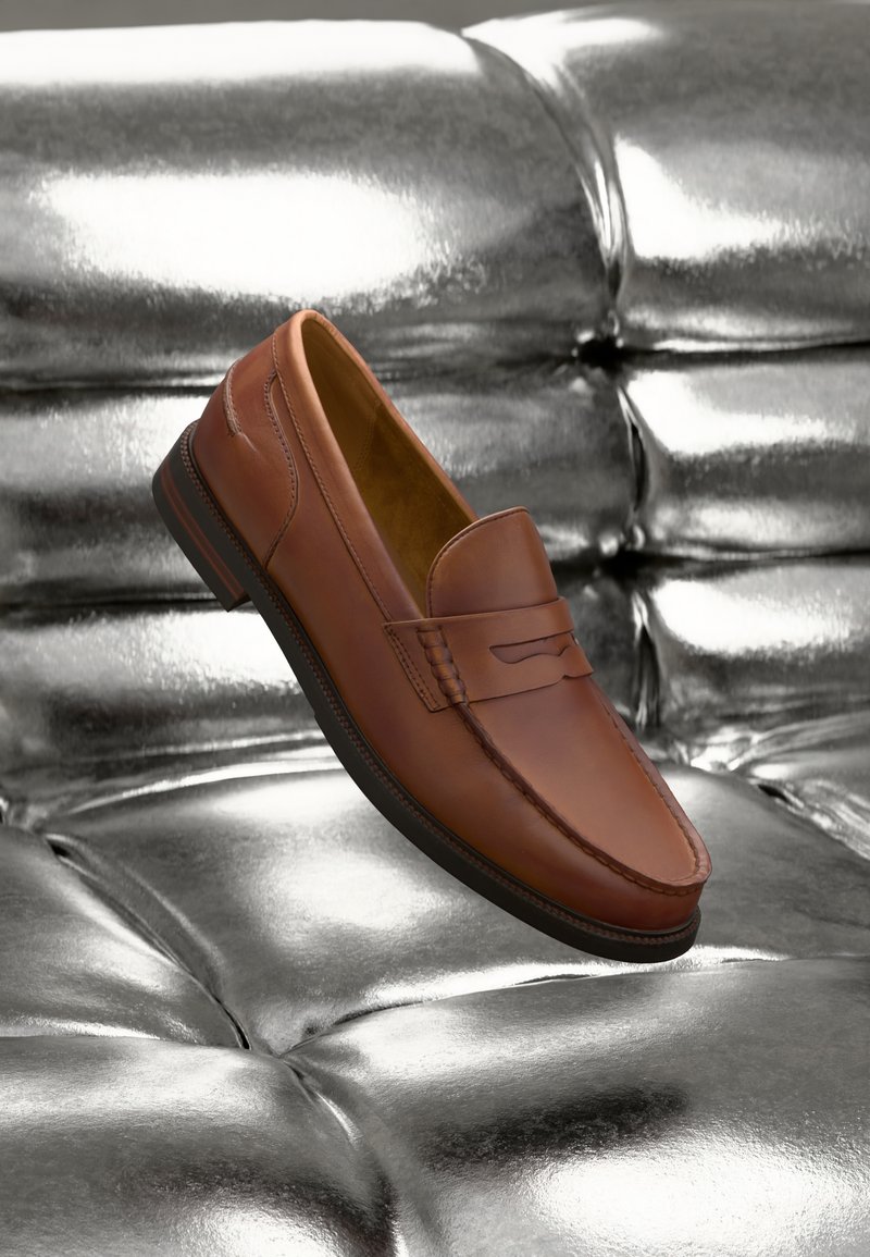 Mocassins en cuir marron avec détails de couture, texture lisse et bout arrondi. Placés sur une surface métallique brillante.