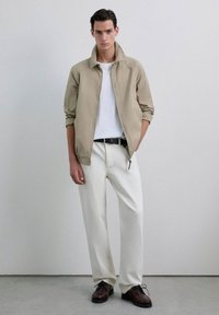 Chaqueta ligera beige con cuello vuelto, camiseta blanca debajo, pantalones crema y zapatos de cordones marrones. Diseño simple y moderno.