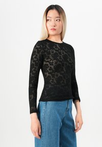 Kvinne med langt rett hår iført en svart blomstret blonde langermet topp og blå høytlivsjeans, som ser til venstre.