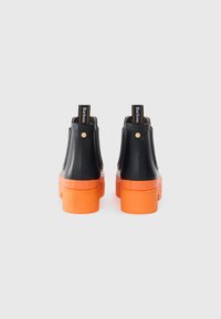 Bottines en caoutchouc noires avec une épaisse semelle orange, dotées de tirants noirs emblématiques et de détails ronds dorés sur le talon.