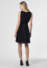 Robe sans manches marine avec un dos plissé, ceinture à la taille et jupe évasée ; présente une texture lisse et un éclat subtil, associée à des talons noirs.