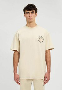 Camiseta de algodón beige con mangas cortas, escote redondo y un parche gráfico circular en la parte frontal. Textura suave y corte relajado.