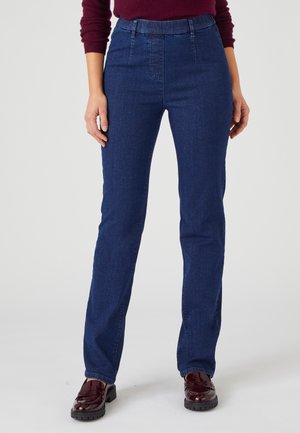 Donna con jeans blu scuro a gamba dritta, top a maniche lunghe color bordeaux e mocassini bordeaux scuro, in piedi su uno sfondo semplice.