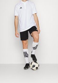 Polo blanc à manches courtes et col, short noir, chaussettes de football noires et blanches rayées, et chaussures de football noires. Ballon de football au sol.