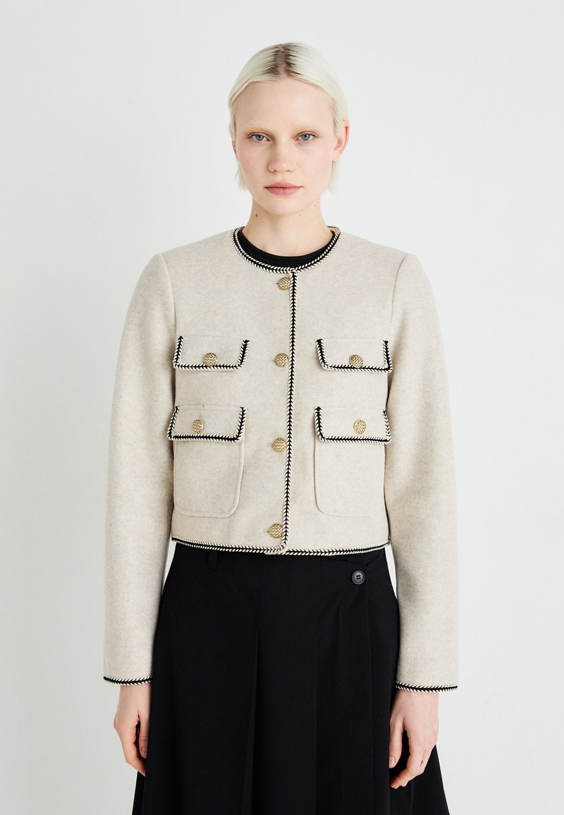 Geknipte beige wollen jas met zwarte rand, voorzien van gouden knopen en twee voortassen, gecombineerd met een zwarte rok. Vlotte textuur, op maat gemaakte pasvorm.