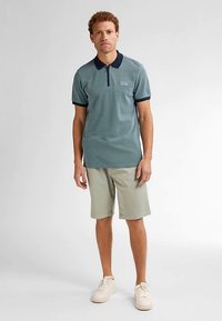 Polokošile s krátkým rukávem v modrozelené barvě s tmavě modrým límcem, texturovaná síťovaná látka a zapínání na zip. Kombinováno s lehkými khaki šortkami a béžovými teniskami.