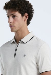 Polo shirt beige à manches courtes en tissu doux, avec une fermeture éclair noire et un bord de col, arborant un petit logo de pingouin sur la poitrine.