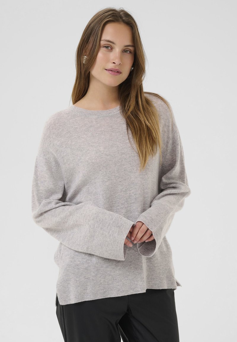 Mlada ženska z dolgimi lasmi, oblečena v svetlo sivo oversized pulover in črne hlače, stoji ob preprosti svetli ozadji.