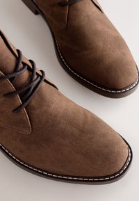 Braune Wildlederschuhe mit rund zulaufender Spitze, dunkel eingefärbten Schnürsenkeln und kontrastierend hellen Nähten entlang der Sohle.