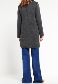 Szary wełniany płaszcz o wykończeniu fakturowanym, z długimi rękawami i prostym dołem, w połączeniu z rozszerzanymi niebieskimi dżinsami. Widok z tyłu.