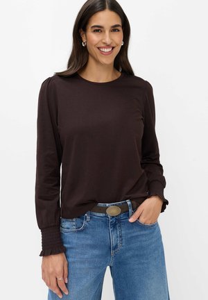 Frau lächelt, trägt ein dunkelbraunes Langarmshirt mit gerafften Bündchen und blaue Jeans mit schwarzem Gürtel und runder Schnalle.
