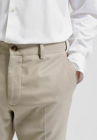 Beige skräddarsydda byxor i strukturerad tyg, med knappstängning, slät framsida och sidofickor. I kombination med en vit skjorta.