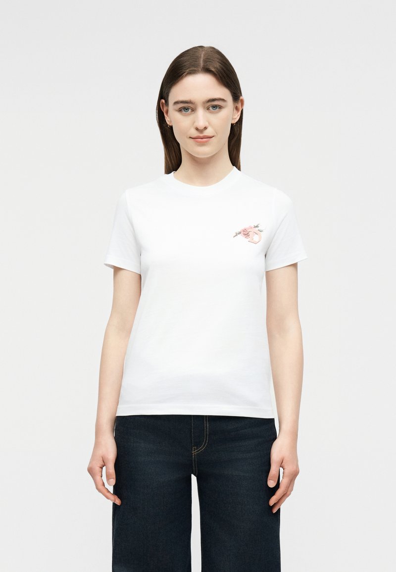 Νεαρή γυναίκα με ίσια καστανά μαλλιά φοράει λευκό κοντομάνικο μπλουζάκι με μικρό λουλουδένιο σχέδιο στο αριστερό στήθος και σκούρο μπλε τζιν.