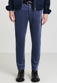 Marineblaue Cordhosen mit schmaler Passform, die eine dezente Textur und Seitentaschen aufweisen, kombiniert mit schwarzen Stiefeletten und einer Jacke mit Reißverschluss.