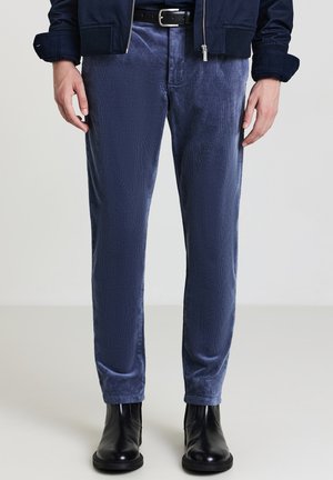 Marineblaue Cordhosen mit schmaler Passform, die eine dezente Textur und Seitentaschen aufweisen, kombiniert mit schwarzen Stiefeletten und einer Jacke mit Reißverschluss.