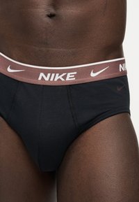 Αρσενικός κορμός που φοράει μαύρα εσώρουχα Nike με καφέ μέση που διαθέτει λευκά λογότυπα και κείμενο Nike.