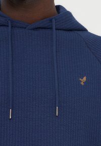 Κοντινό πλάνο ενός ατόμου που φοράει υφασμάτινο σκούρο μπλε φούτερ με κορδόνια και ένα μικρό κεντητό χρυσό πουλί στο στήθος.