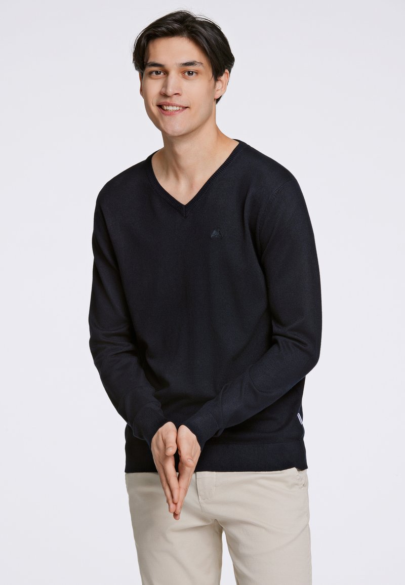 Schwarzer V-Ausschnitt-Pullover aus glattem Stoff, mit langen Ärmeln und einem gerippten Saum. Das Modell trägt hellbeige Hosen.