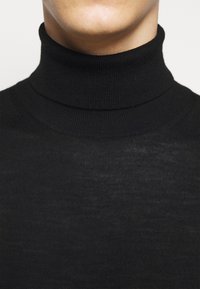 Gros plan sur une personne portant un pull à col roulé noir côtelé, montrant le cou et la partie supérieure de la poitrine.