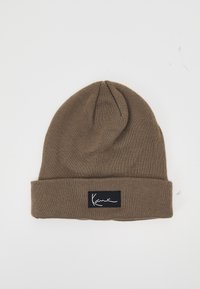Brown knit beanie with a folded cuff, featuring a black label with white text. Soft texture; rounded top tapering slightly.