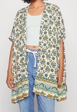 Femme portant un kimono crème ouvert avec des motifs floraux bleus et dorés, associé à un crop top bleu clair et un jean en denim à cordon.