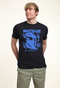 Černé tri�čko s modrou grafickou potiskem obsahující text "MATISSE" a design z "Blue Nude II" (1952), umístěné na neutrálním pozadí.