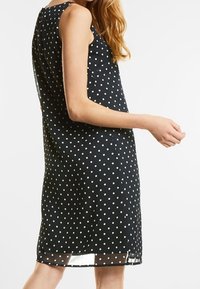 Svart ärmlös klänning med vita prickar, rak silhuett och genomskinlig fåll. Tillverkad av lätt material, lämplig för varmt väder.