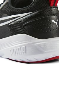 Zapatilla deportiva negra con parte superior de malla texturizada y cuero sintético, que presenta rayas blancas y un acento rojo en el talón, y una suela blanca.