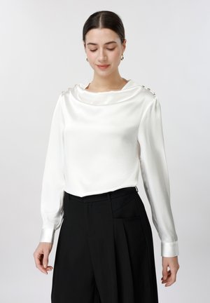 Γυναίκα με σκοτεινά μαλλιά, φορεμένη με λευκό μακρυμάνικο πουκάμισο με κουμπιά στους ώμους και μαύρο παντελόνι ψηλόμεσο, κοιτάει προς τα κάτω με ελαφρύ χαμόγελο.