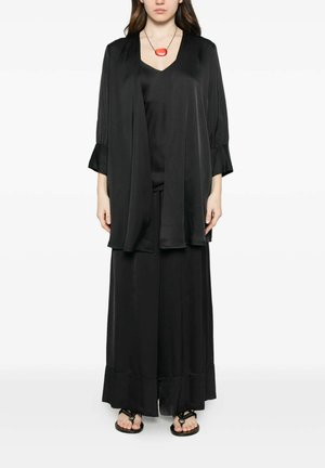 Donna che indossa un outfit largo in raso nero con pantaloni ampi e giacca lunga aperta sul davanti, sandali neri e una collana con ciondolo ovale rosso.