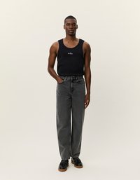 Melnā bezplecu blūze ar logo, ko kombinē ar augstām, brīvām pelēkām džinsa biksēm un melnām apaviem. Neitrāla fonu uzsver uzmanību uz apģērbu.