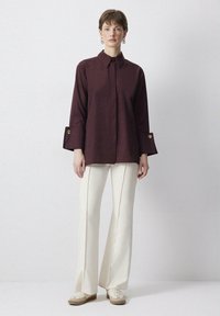 Camicia button-up color bordeaux con maniche lunghe e bottoni dorati, abbinata a pantaloni larghi color crema e sneakers beige.