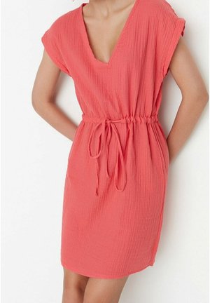 Femme portant une robe courte corail avec un décolleté en V et une taille à cordon, debout les mains derrière le dos sur un fond blanc.