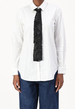 Persoon draagt een wit overhemd met lange mouwen en donkerblauwe spijkerbroek, met een smalle zwarte sjaal met bloemenkant om de hals gestrikt.