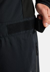 Czarna kurtka outdoorowa z gładkiego materiału, z siateczkowym panelem, regulowanym paskiem na rzep oraz detalem kieszeni na zamek.