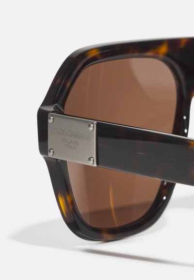 Ochelari de soare cu nuanță maronie, cu un cadru din tortoise, având o placă de metal accentuată, gravată cu "Dolce & Gabbana Milano Italia."
