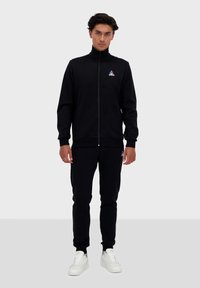 Tuta sportiva nera realizzata in un tessuto morbido, caratterizzata da una giacca con zip e pantaloni abbinati. Accenti di sneaker bianchi completano il look.