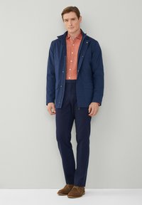 Chaqueta azul marino con un cuello alto, llevada sobre una camisa de botones color coral y pantalones azul marino. Zapatos de ante marrón completan el conjunto.